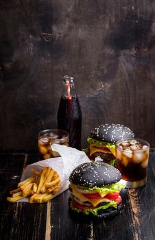 Set of black burgers with meat patty, cheese, tomatoes, mayonnaise, french fries in a paper cup and glass of cold cola soda with ice. Dark wooden rustic table. Modern fast food lunch
