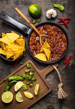 Chili con carne in frying pan on dark wooden background. Ingredients for making Chili con carne. Top view. Chili with meat, nachos, lime, hot pepper. Mexican/Texas traditional dish Chili con carne