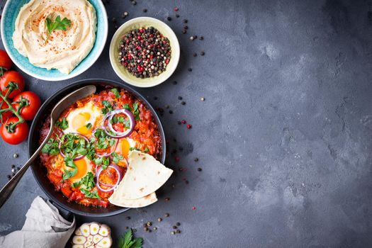Shakshuka with pita bread in pan, hummus in bowl on rustic background. Middle eastern traditional dishes. Fried eggs with vegetables. Top view. Space for text. Middle eastern style breakfast or lunch