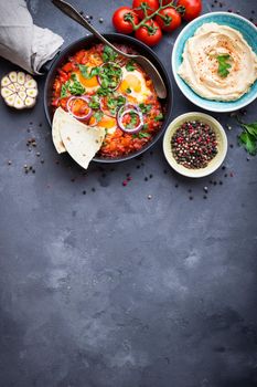 Shakshuka with pita bread in pan, hummus in bowl on rustic background. Middle eastern traditional dishes. Fried eggs with vegetables. Top view. Space for text. Middle eastern style breakfast or lunch