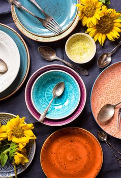 Vintage multicolored empty plates and bowls on a dark gray linen tablecloth with sunflower. With antique spoons and forks. Table setting. Shabby chic/retro style. Top view. Rustic kitchen