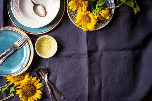 Vintage multicolored empty plates and bowls on a dark gray linen tablecloth with sunflower. With antique spoons and forks. Space for text. Table setting. Shabby chic/retro style. Top view. Copy space