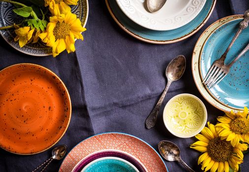 Vintage multicolored empty plates and bowls on a dark gray linen tablecloth with sunflower. With antique spoons and forks. Space for text. Table setting. Shabby chic/retro style. Top view. Copy space