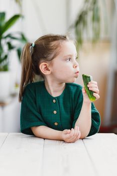 Cute little girl in green muslin dress eating cucumber, fresh vegetables, healthy food concept, indoor portrait.