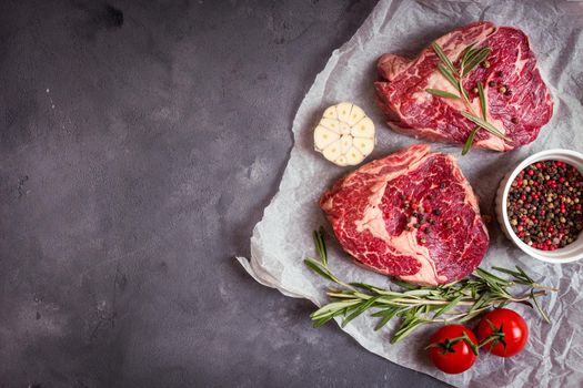 Raw juicy ribeye steaks with seasonings ready for roasting on baking paper. Rustic concrete background. Fresh marbled meat steaks with herbs, garlic, oil, spices, tomatoes. Space for text. Top view