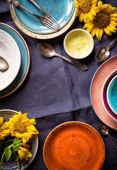 Vintage multicolored empty plates and bowls on a dark gray linen tablecloth with sunflower. With antique spoons and forks. Space for text. Table setting. Shabby chic/retro style. Top view. Copy space