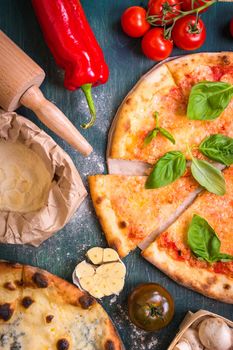 Delicious sliced pizza and ingredients for making pizza. Flour, cheese, tomatoes, basil, pepperoni, mushrooms and rolling pin over wooden background. Top view. Pizza ready to eat