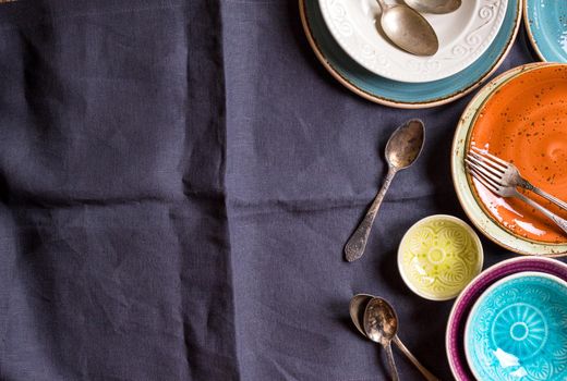 Vintage multicolored empty plates and bowls on a gray linen tablecloth. With antique spoons and forks. Space for text. Table setting. Shabby chic/retro style. Top view. Rustic kitchen. Copy space