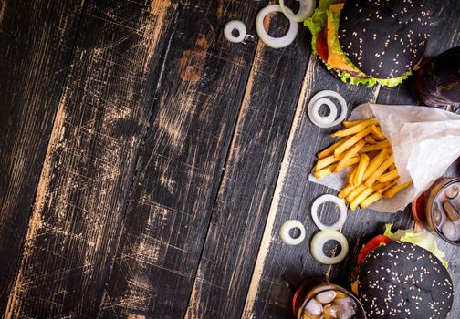Set of black burgers with meat patty, cheese, tomatoes, mayonnaise, french fries and glass of cold cola soda with ice from above. Dark wooden rustic background. Space for text. Top view