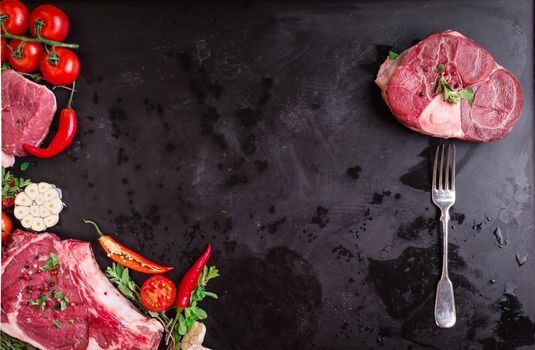 Raw juicy meat steaks ready for roasting on a black chalk board background. Meat on a fork. Veal shank (ossobuco), rib eye steak on the bone, fillet with cherry tomatoes, hot pepper and herbs.