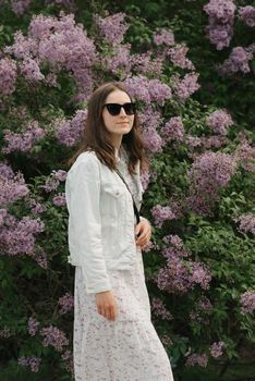 A woman with long hair is staying near the lilac bush in the village green. A lady in black sunglasses is posing in the woodland.