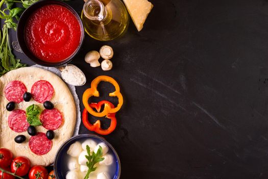 Pizza making background. Ingredients for making pizza. Space for text. Pizza dough, flour, cheese, mozzarella, tomatoes, basil, pepperoni, olives and rolling pin over black chalkboard. Top view