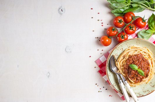 Italian pasta bolognese. Spaghetti with meat and tomato sauce in a plate with Italian tablecloth on a wooden white background. With fresh cherry tomatoes, basil. Food frame. Space for text. Top view