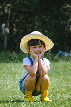 Little girl in a hat makes cute poses and smiles at the camera sitting in the grass on a bright summer day. Cute girl playing in a beautiful garden. A little gardener.