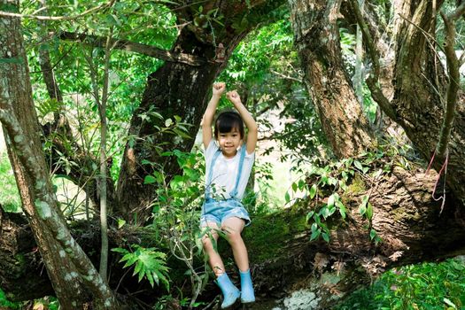 Cute little girl in boots playing in the garden, climbing a tree. Happy girl relaxing on tree branch.