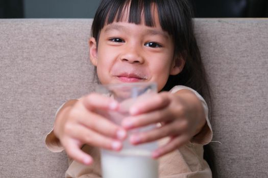 Portrait of a cute Asian little girl holding a glass of milk sitting on the sofa at home. Small girl at home with smiling face, feeling happy enjoying drinking milk and looking at camera.