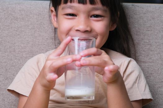 Portrait of a cute Asian little girl holding a glass of milk sitting on the sofa at home. Small girl at home with smiling face, feeling happy enjoying drinking milk and looking at camera.