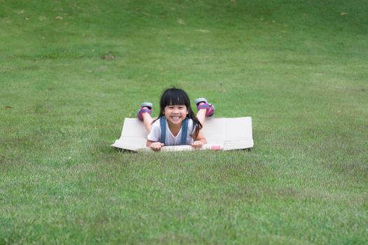 Smiling little girl lies prone on a cardboard box sliding down a hill at a botanical garden. The famous outdoor learning center of Mae Moh Mine Park, Lampang, Thailand. Happy childhood concept.
