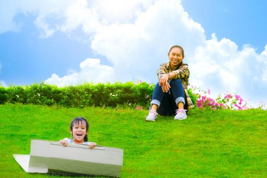 Smiling little girl sat on a cardboard box sliding down a hill at the Botanical Garden and her mother smiled as she looked at her. Happy childhood concept.