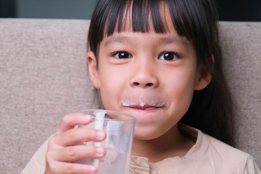 Portrait of a cute Asian little girl holding a glass of milk sitting on the sofa at home. Small girl at home with smiling face, feeling happy enjoying drinking milk and looking at camera.