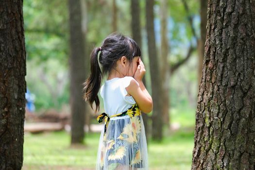 Smiling little girl covering her eyes with both hands, playing hide and seek standing beside a large tree. Cute little girl having fun outdoors in the park.