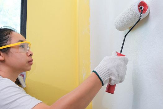Young woman decorator wearing protective goggles paints the walls with a roller. Repair and interior decoration.