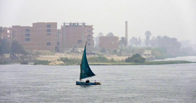 A dhow with a blue sail sailing over the Nile. A dhow is a traditional wooden sailing boat. In the distance is the vague outline of an Egyptian town visible
