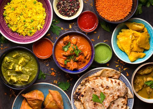 Assorted indian food on dark wooden background. Dishes and appetizers of indian cuisine. Curry, butter chicken, rice, lentils, paneer, samosa, naan, chutney, spices. Bowls and plates with indian food