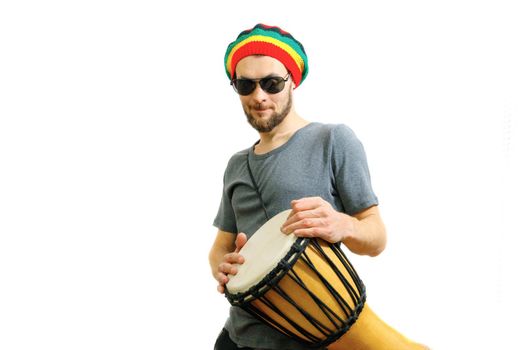 Young caucasian smiling man in rasta hat, sunglasses and grey t-shirt on white background with djembe african drum