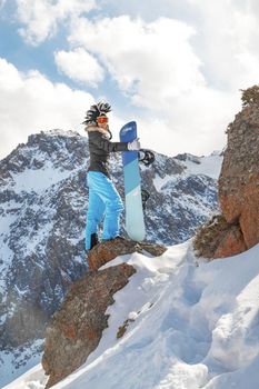 Snowboarder woman in mohawk hat on top of rock in snowy mountain. Copy space