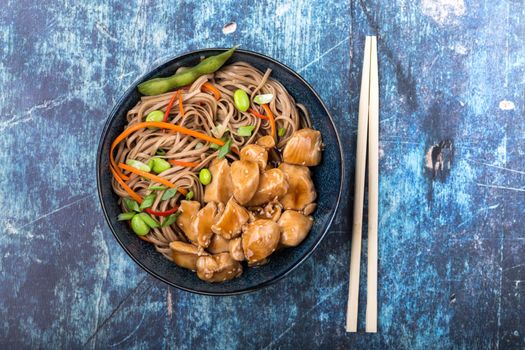 Asian noodles with chicken, vegetables, bowl, rustic wooden blue background. Closeup. Top view. Soba noodles, teriyaki chicken, edamame, chopsticks. Asian style dinner/lunch. Chinese/Japanese noodles