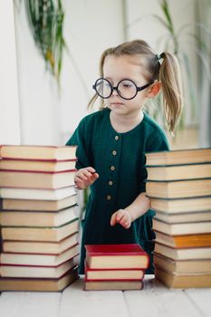 Cute little girl in round shaped glasses and green muslin dress folds colorful books in order.