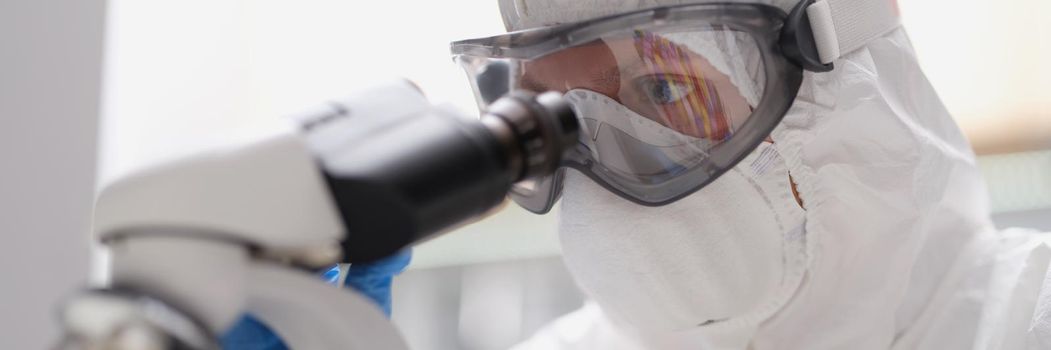 A man in a protective suit and mask looks through a microscope, close-up. Dangerous virus research laboratory
