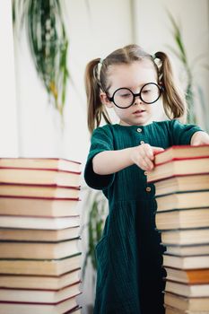 Little cute preschool age girl in round shaped glasses between books pile.