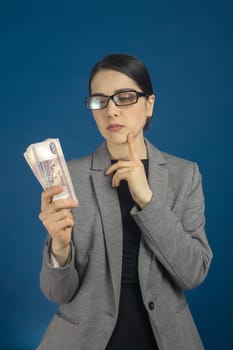 Young woman in glasses looks at a pack of Russian ruble with interest on blue background. Vertical, selective focus.