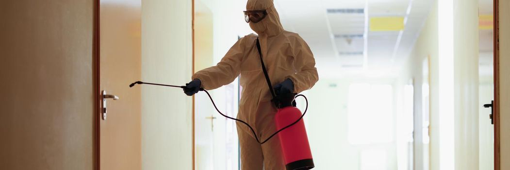 A man in a protective suit splashes on the door, the corridor of the room. Antiviral protection during the covid pandemic