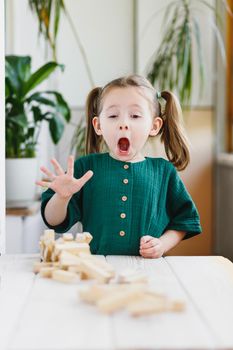 Preschool age girl in green dress expressive reaction on unexpected falling of wooden block tower.