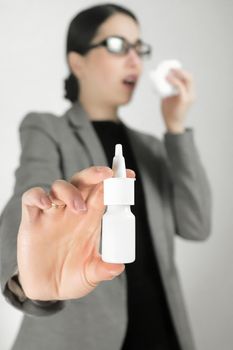 Young beautiful woman in glasses and gray jacket holding nose spray in front of her. Selective focus, vertical.