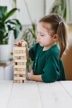 Cute little girl in green dress putting a wooden block on top of jenga tower.