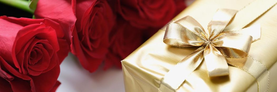 Red rose buds and a gift in gold paper with a ribbon, close-up. Template for greeting card, present event