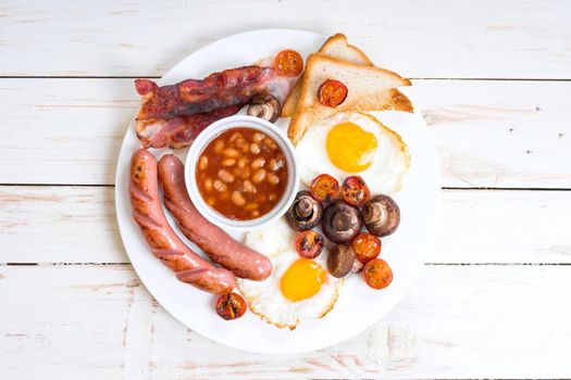 Full english breakfast with fried eggs, tomatoes, sausages, bacon, mushrooms, toasts and beans. Breakfast on a white plate with forks on the white wooden table. Top view