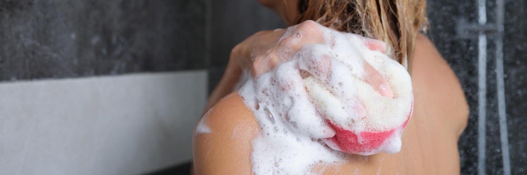 Woman washes her body with a sponge with shower gel, close-up rear view. Soap foam on a washcloth