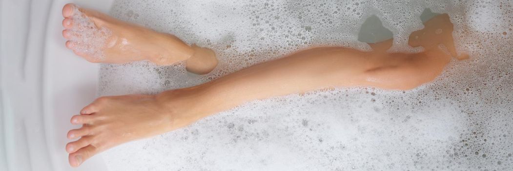 Slender female legs in a bathtub with soapy water, close-up. Bath foam, home aromatherapy, bath salt