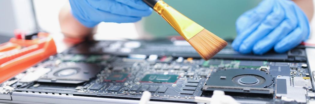 A man in gloves with a brush cleans the details of a laptop, close-up. Dust cleaning, partial disassembly of the computer