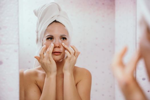 Young beautiful woman using a moisturizing facial mask after taking a bath. Pretty attractive girl in a towel on her head stands in front of a mirror in a home bathroom. Daily hygiene and skin care.