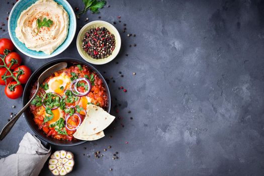 Shakshuka with pita bread in pan, hummus in bowl on rustic background. Middle eastern traditional dishes. Fried eggs with vegetables. Top view. Space for text. Middle eastern style breakfast or lunch