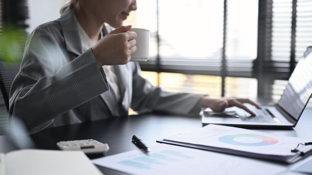 Professional businesswoman holding coffee cup and working with laptop computer at office.