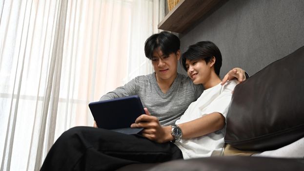 Happy gay couple using digital tablet while spending leisure time together at home. Concept of sexual freedom and equal rights for LGBT community.