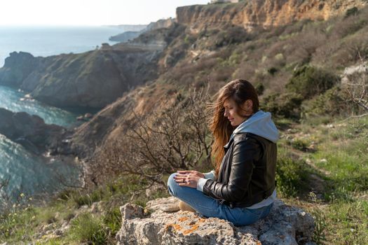 Woman tourist enjoying the sunset over the sea mountain landscape. Sits outdoors on a rock above the sea. She is wearing jeans, a blue hoodie and a black leather jacket. Healthy lifestyle, harmony and meditation.