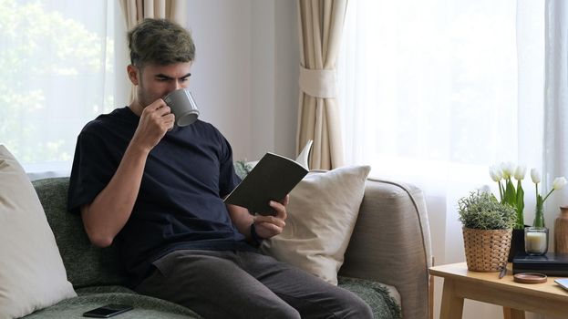 Relaxed millennial man drinking hot coffee and reading book on comfortable sofa.